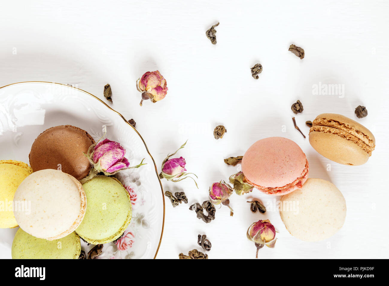 Multicoloured macaroons on beautiful porcelain plate, tea and dried rose buds on white background; top view, flat lay Stock Photo