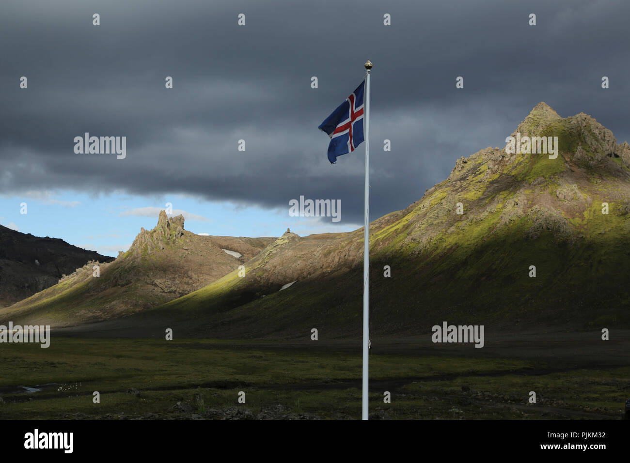 Iceland, Icelandic flag blowing in front of dramatic landscape, evening light, black clouds and sunshine Stock Photo