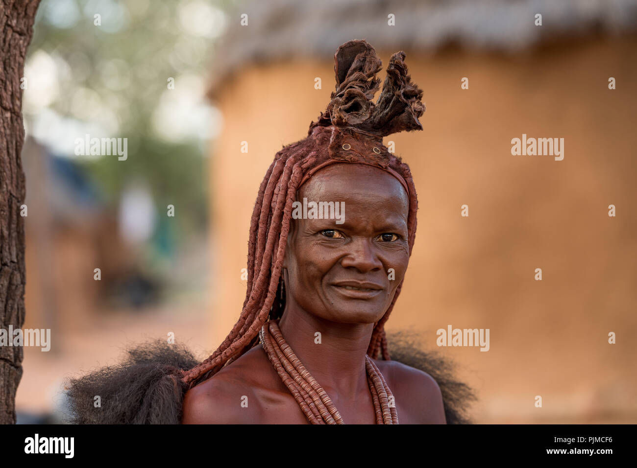 Himba woman smiles and looks into the camera. Leather crown and hair pots are very visible Stock Photo