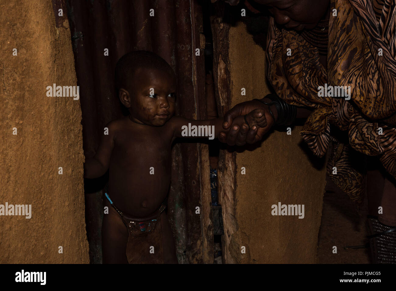 Himba boy with traditional loincloth stands in the dark in the entrance of a mud hut on the hand of his mother Stock Photo
