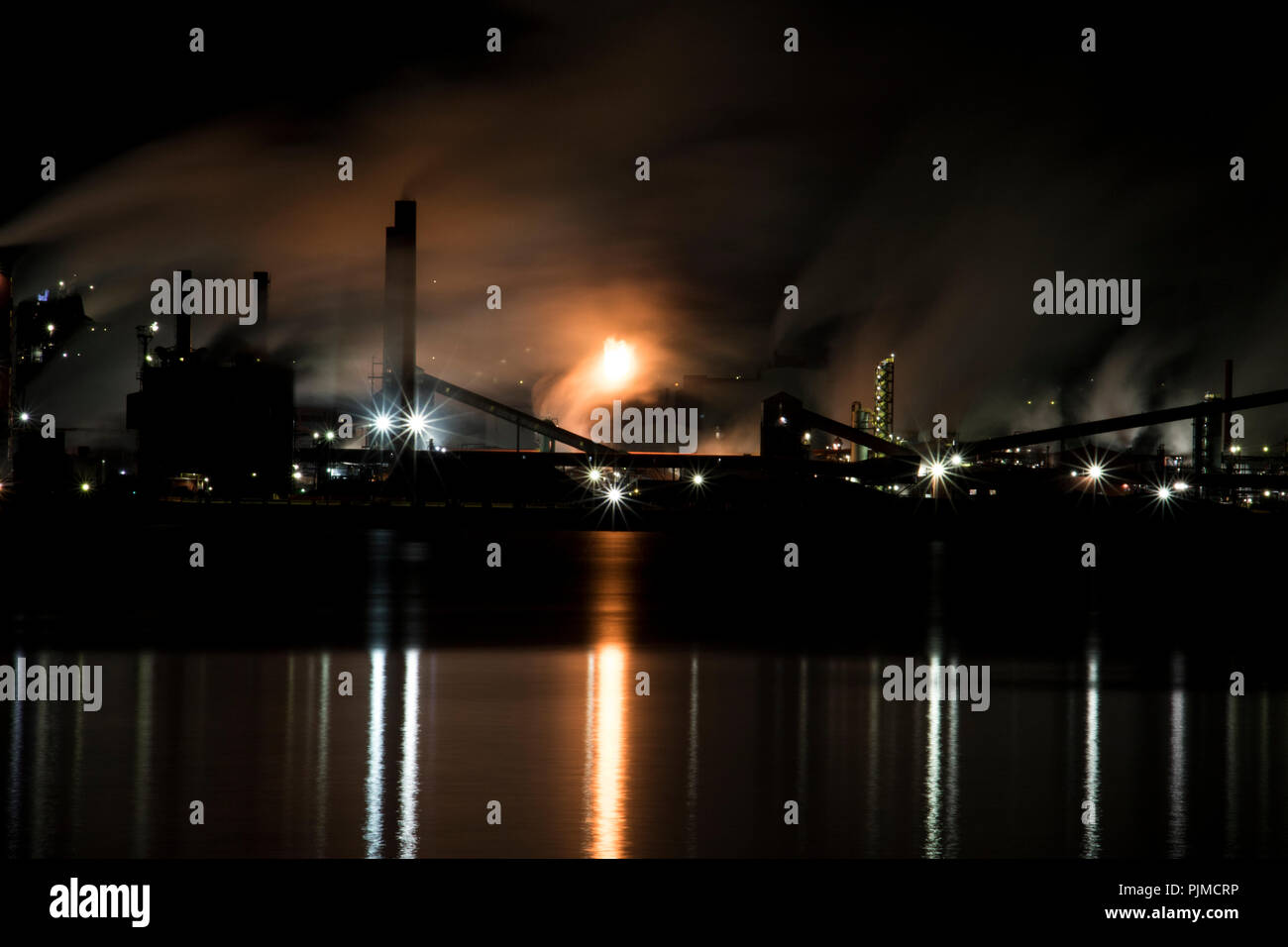 Industrial steel mill at night in the city over looking water. Smoke blowing in the wind flowing out of factory smoke stacks creating air pollution. G Stock Photo
