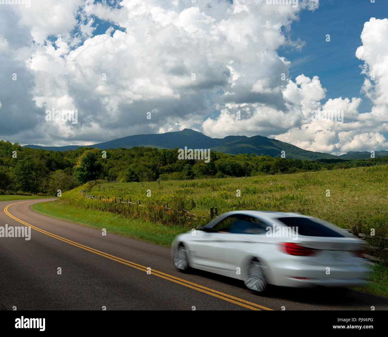 Car Driving on Blue Ridge Parkway North Carolina - Grandfather Mountain in distance as a car drives by fields of yellow flowers and blue clouds skies. Stock Photo