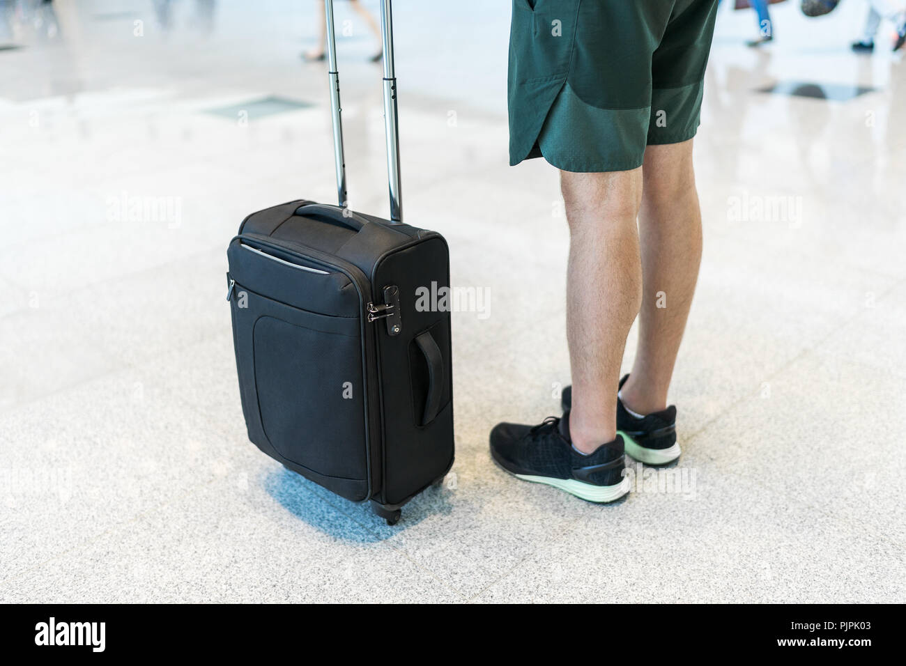 Young man wearing shorts and sneakers is standing at the airport holding a  small-sized black suitcase with 4 wheels Stock Photo - Alamy