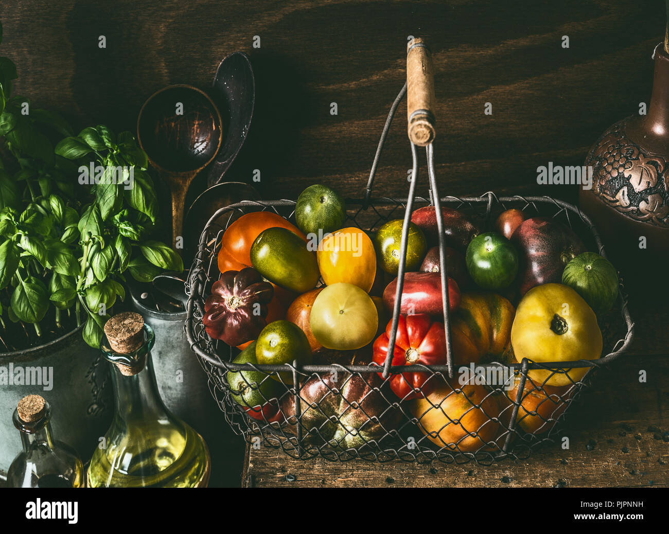 Colorful organic tomatoes in harvest basket on dark rustic kitchen table with cooking ingredients. Healthy food concept Stock Photo