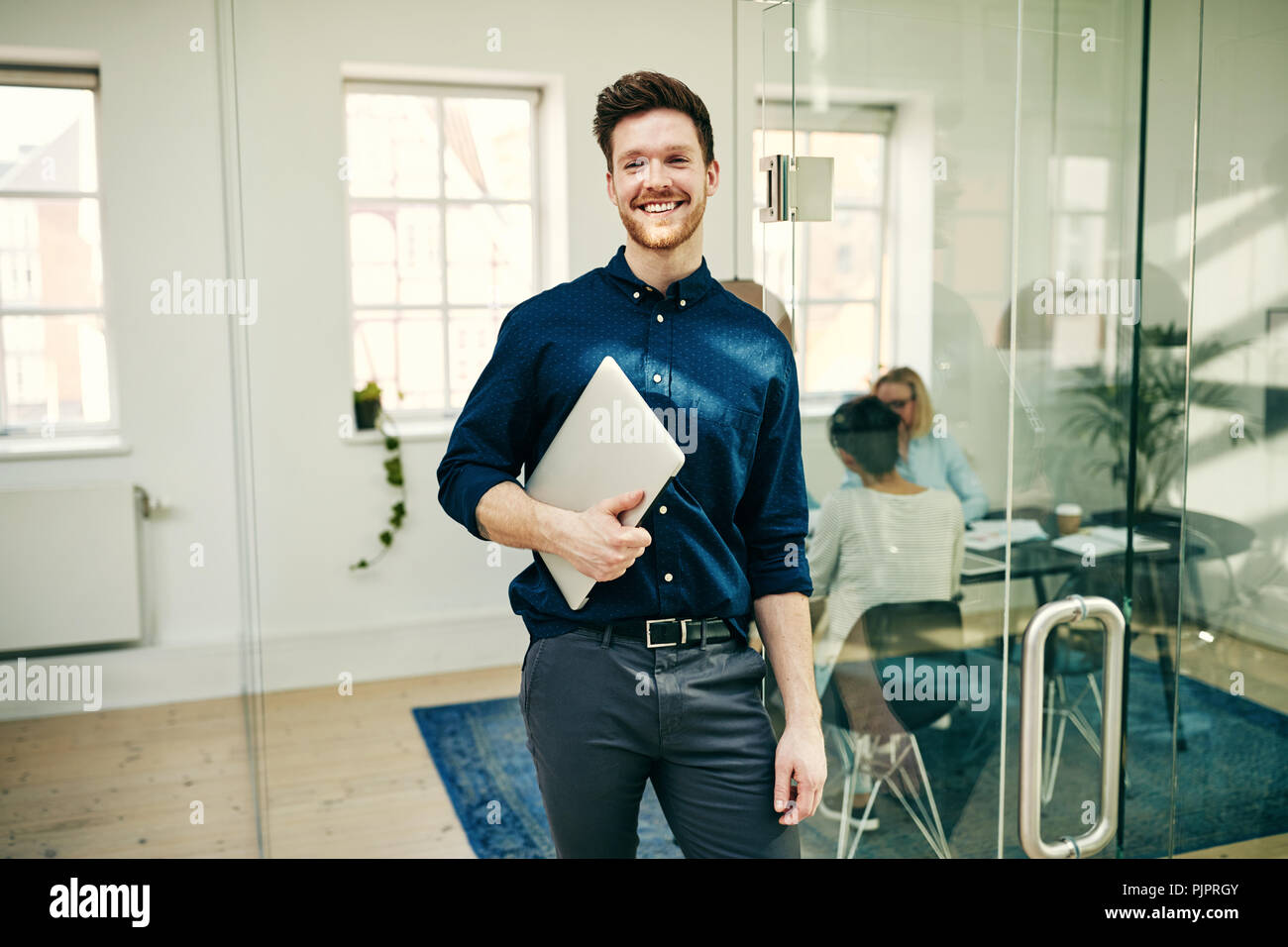 Young businessman smiling while standing in a modern office holding his laptop with colleagues at work in the background Stock Photo