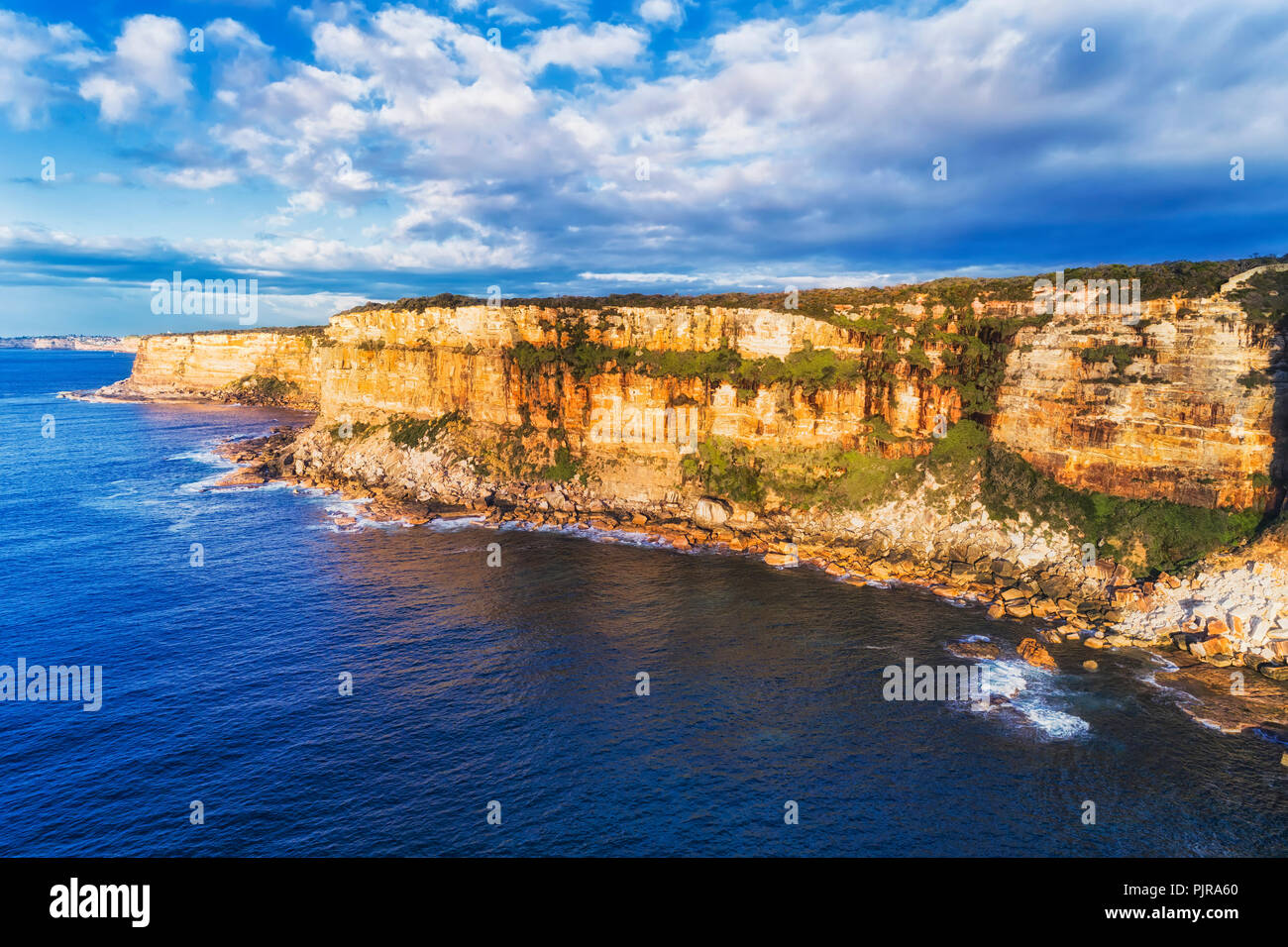 Tall eroded rugged sandstone cliffs of Sydney's North head at the entrance to Sydney harbour hit by endless waves of Pacific ocean lit by warm morning Stock Photo