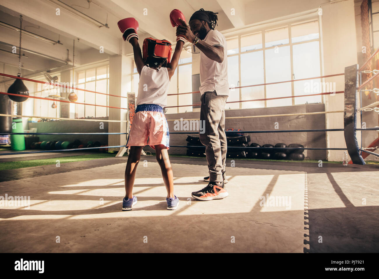 Rear view of a kid standing inside a boxing ring with raised hands along with his trainer. Trainer raising the hand of a boxing kid standing inside a Stock Photo