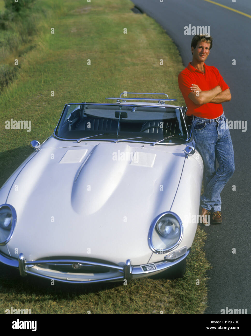 1990 HISTORICAL MAN STANDING ALONE ON ROADSIDE GRASS BY 1966 SERIES 1 XK-E E-TYPE JAGUAR (©JAGUAR CARS LTD 1961) WHITE CONVERTIBLE CAR Stock Photo