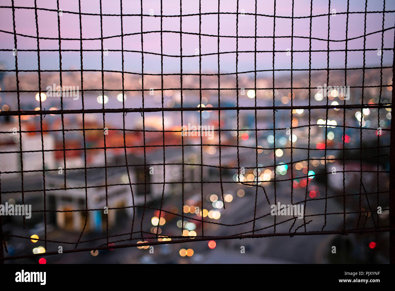 Blurred cityscape of Valparaiso during dusk through a wire fence. Urban abstract, bokeh lights, travel concept, wire fence patterns. Valparaiso, Chile Stock Photo