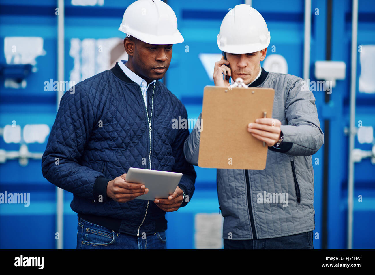 Freight foreman talking on a cellphone and reading an inventory list on a clipboard while standing with a colleague in a commercial shipping yard Stock Photo