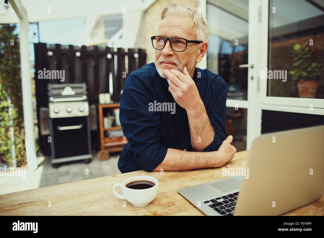 Senior man deep in thought while sitting at a table outside on his patio working on a laptop and drinking a cup of coffee Stock Photo