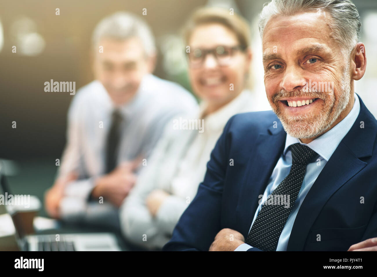 Mature businessman smiling confidently while sitting at a table in an modern office with colleagues in the background Stock Photo