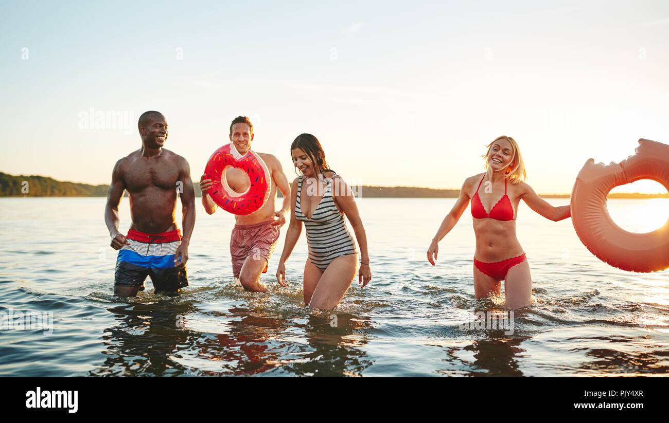 Diveres group of young friends in swimsuits standing in a lake at sunset laughing and splashing water at each other Stock Photo