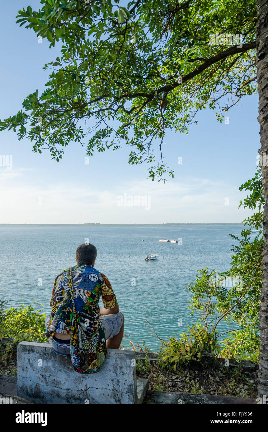 Rear view of young man sitting at coast overlooking the ocean wearing colorful african shirt, Bubaque, Bijagos Islands, Guinea Bissau Stock Photo