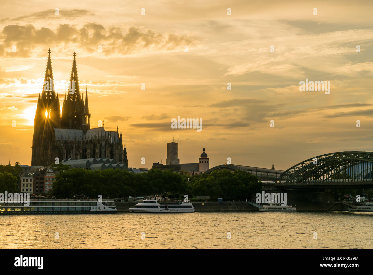 Sunset behind the twin gothic spires of Cologne Cathedral along side the Hohenzollern Bridge and river cruise boats on the River Rhine, Germany, Europ Stock Photo