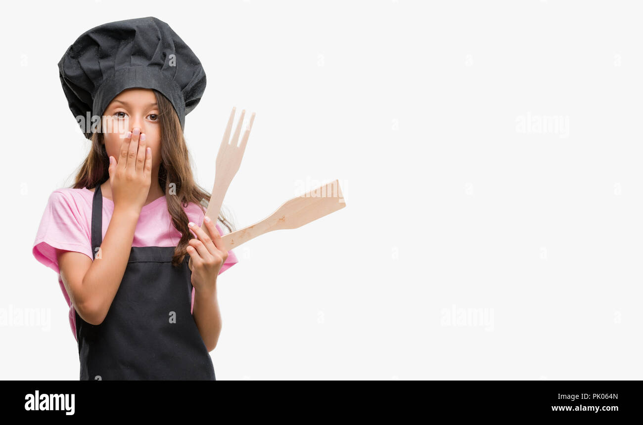 Brunette hispanic girl wearing cook uniform cover mouth with hand shocked with shame for mistake, expression of fear, scared in silence, secret concep Stock Photo