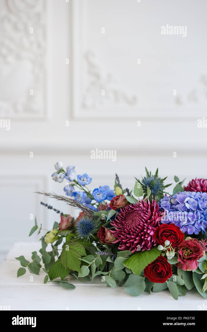 Elongated floral arrangement in vintage metal vase. table setting. Blue and red color. Gorgeous bouquet of different flowers. Bright room, stucco on the wall Stock Photo