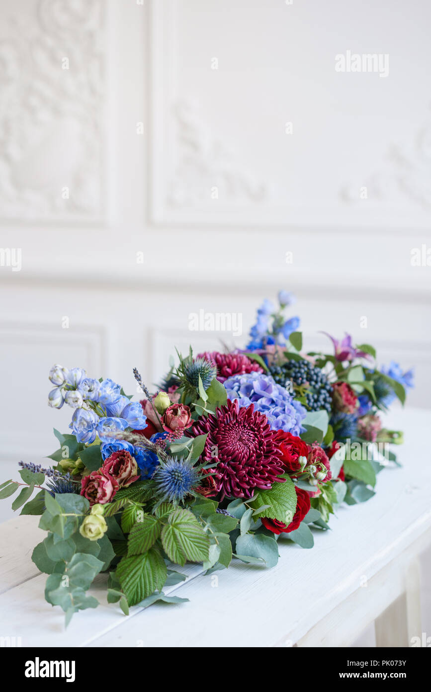 Elongated floral arrangement in vintage metal vase. table setting. Blue and red color. Gorgeous bouquet of different flowers. Bright room, stucco on the wall Stock Photo