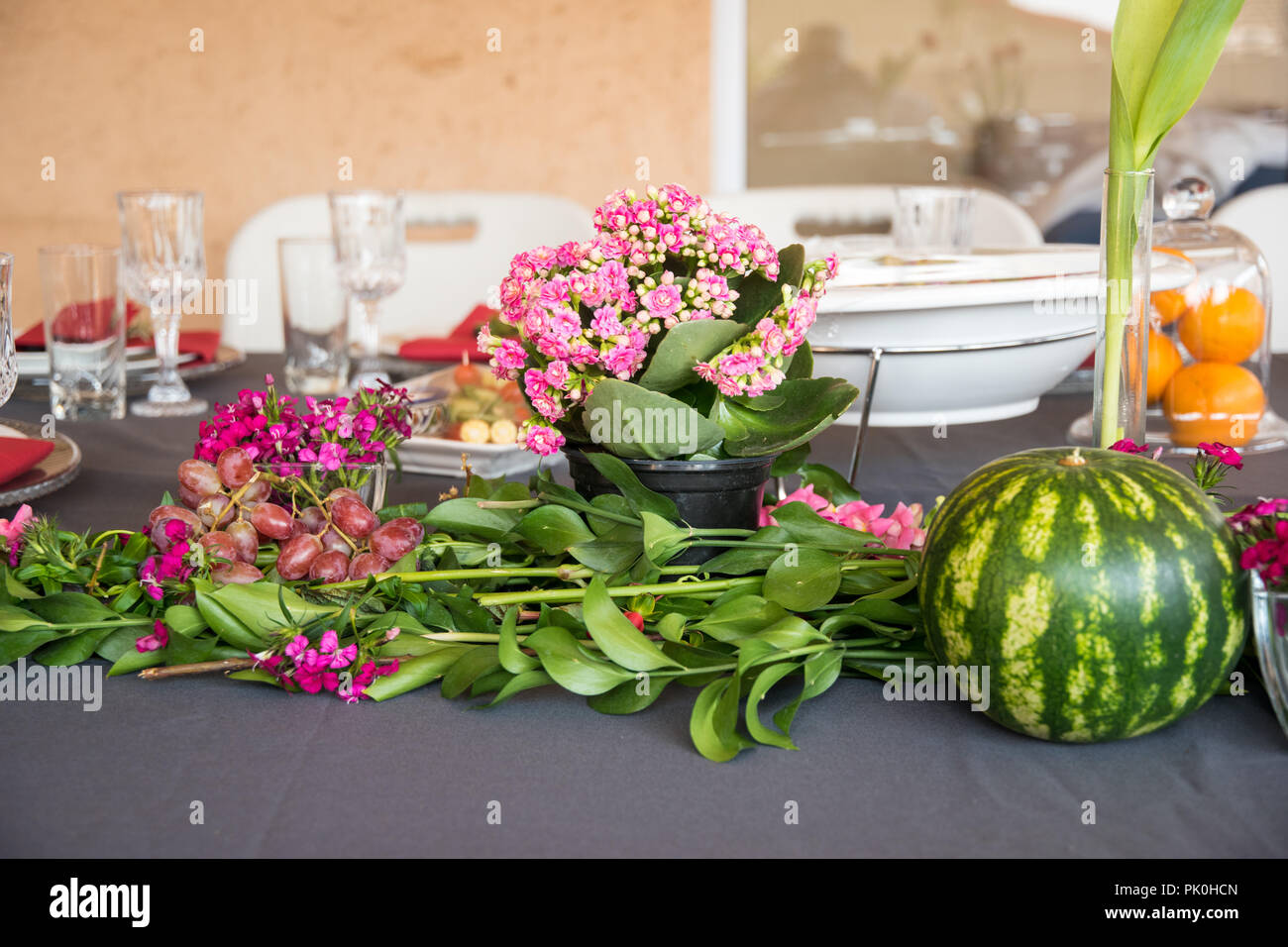 A lunch table with floral display of lovely orange flowers with colourful pink and reddish arrangements in foreground with a round watermelon & grapes Stock Photo
