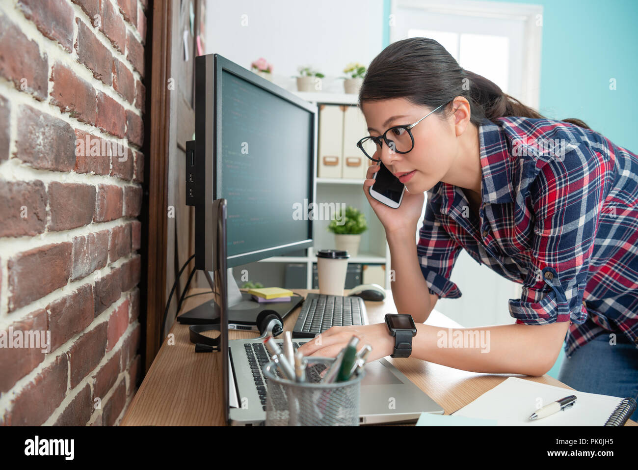 young programmer checking information on laptop and replying her costumer by smartphone. Stock Photo