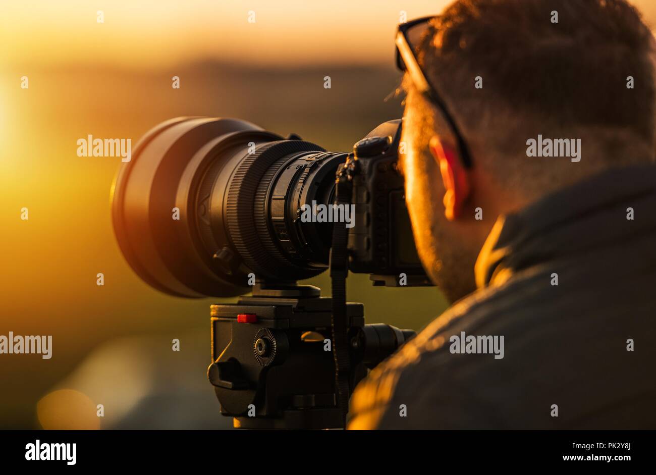 Outdoor Telephoto Photography. Caucasian Photographer in His 20s with Large Telephoto Lens Taking Pictures During Scenic Sunset Using Camera Tripod. Stock Photo