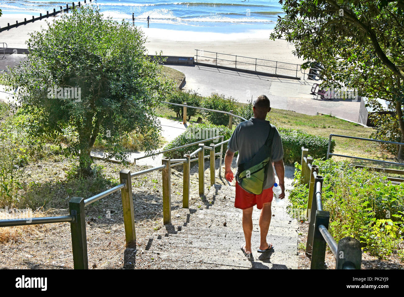 Back view of man in red shorts walking down steps towards early morning people on sandy family seaside holiday beach Frinton Essex coast England UK Stock Photo