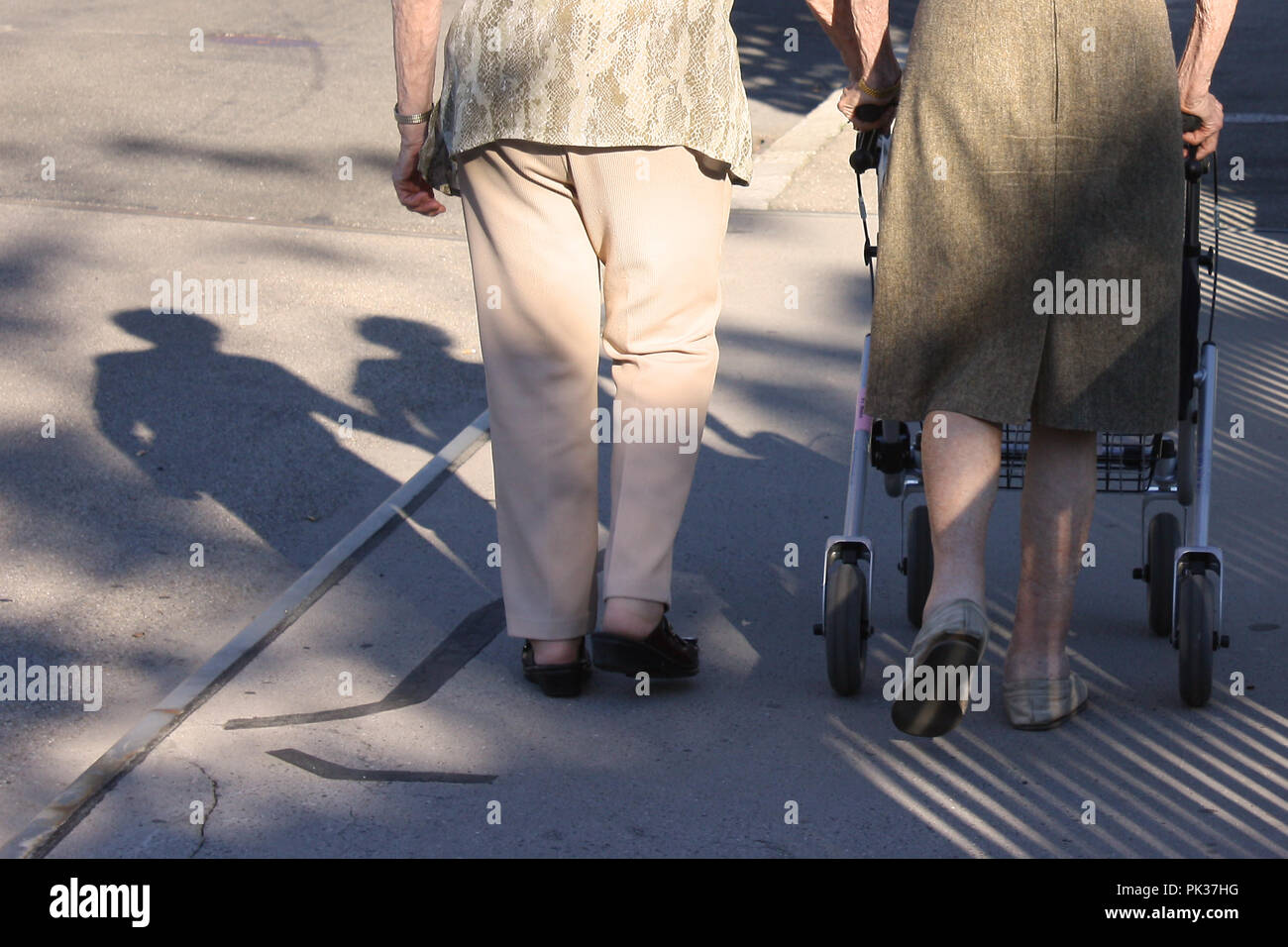 Two old women seen from the back, shadow on the street, walking slowly with a walking frame, iconic photo Stock Photo