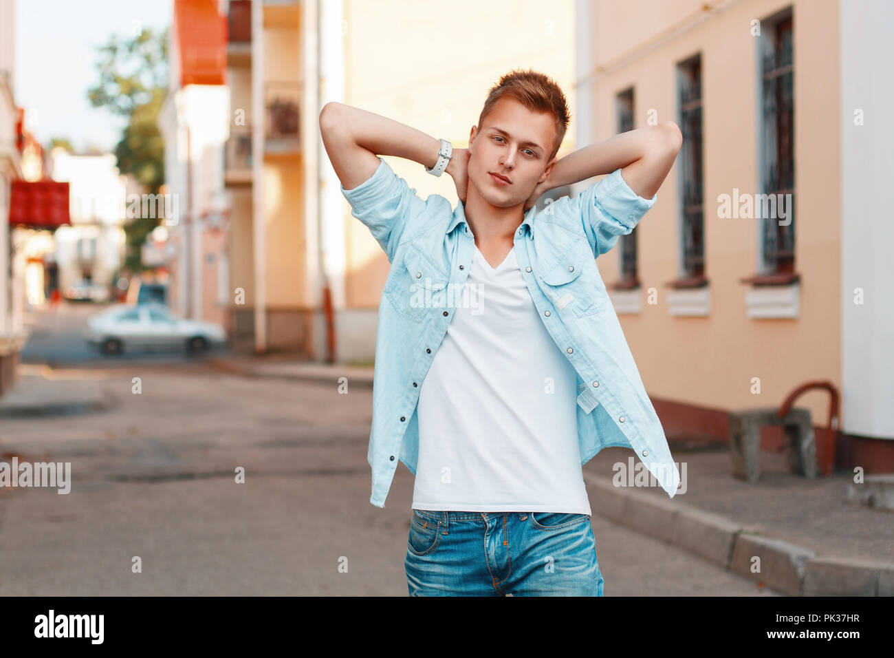 Young handsome man in a denim shirt and jeans holding hands behind head on the background of the city. Stock Photo