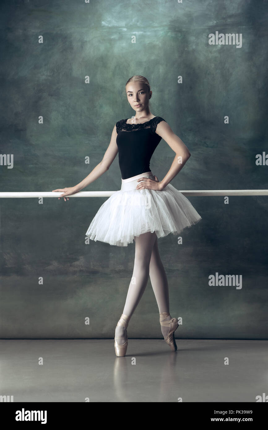 The classic ballet dancer in white tutu posing at ballet barre on studio  background. Young teen