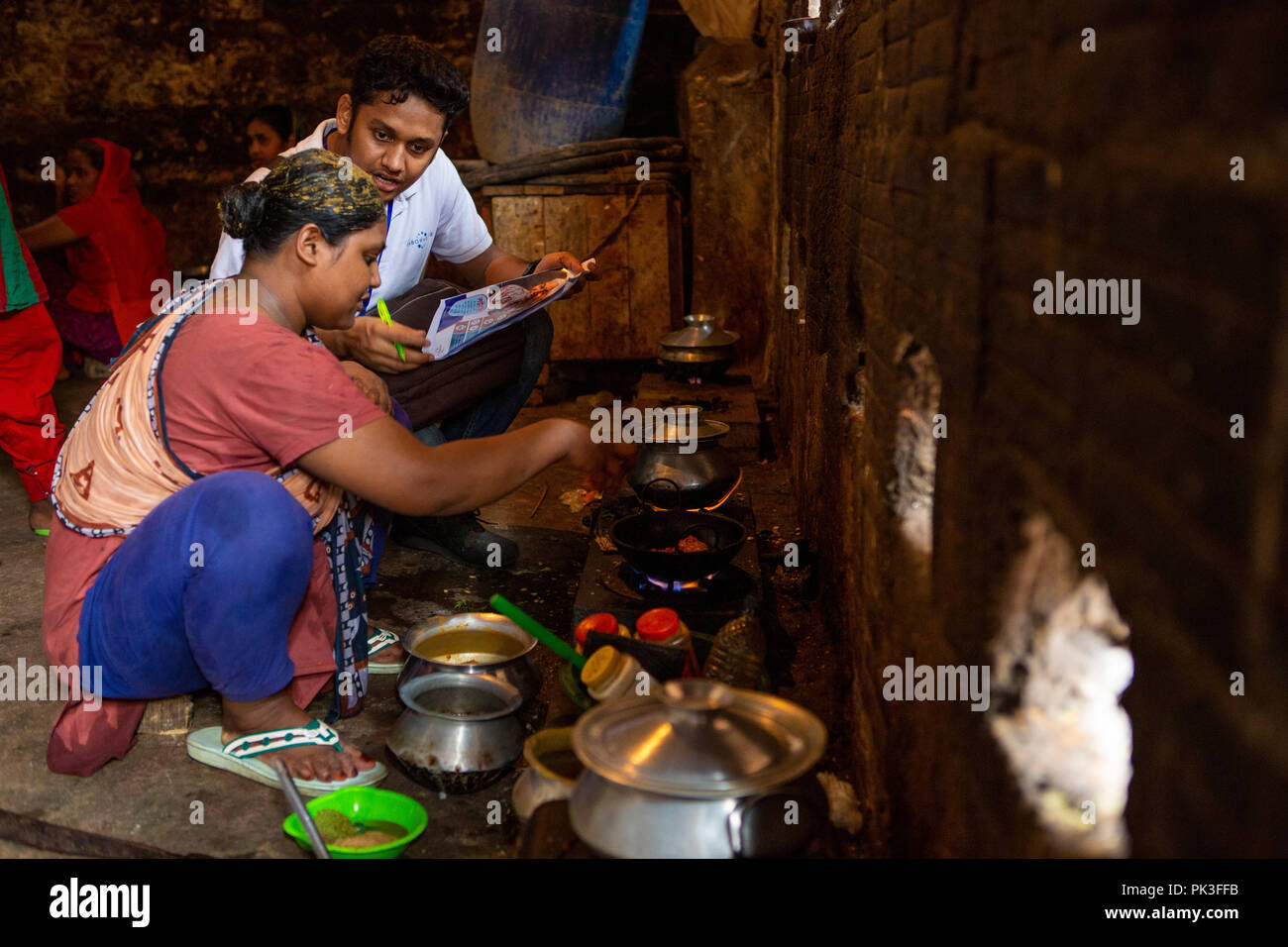 A man talking to a woman about the working conditions of her garment factory as she cooks in a communal kitchen in the slums of Dhaka, Bangladesh. Stock Photo