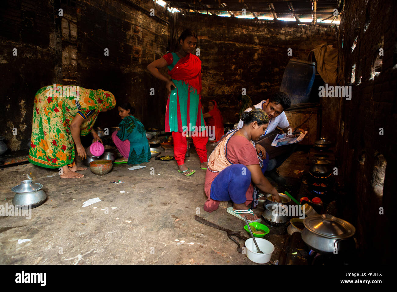 A man talking to a woman about the working conditions of her garment factory as she cooks in a communal kitchen in the slums of Dhaka, Bangladesh. Stock Photo