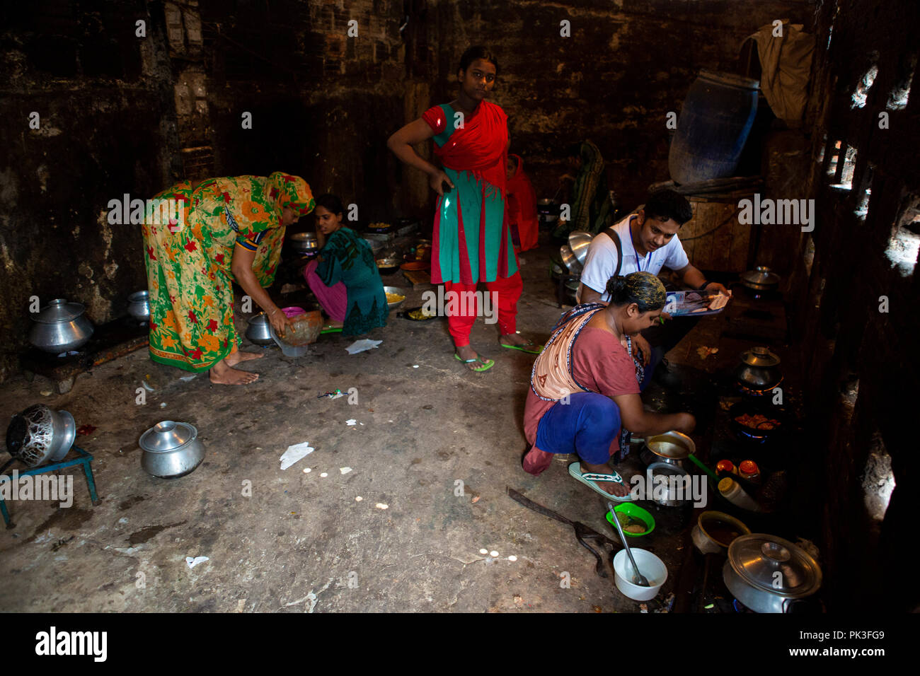 A man talking to a woman about the working conditions of her garment factory as she cooks in a communal kitchen in the slums of Dhaka, Bangladesh. Stock Photo