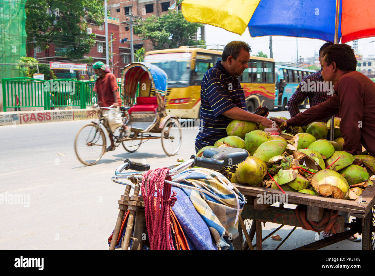A street vendor selling fresh coconuts as a cycle rickshaw passes in the background on the street in Dhaka, Bangladesh. Stock Photo
