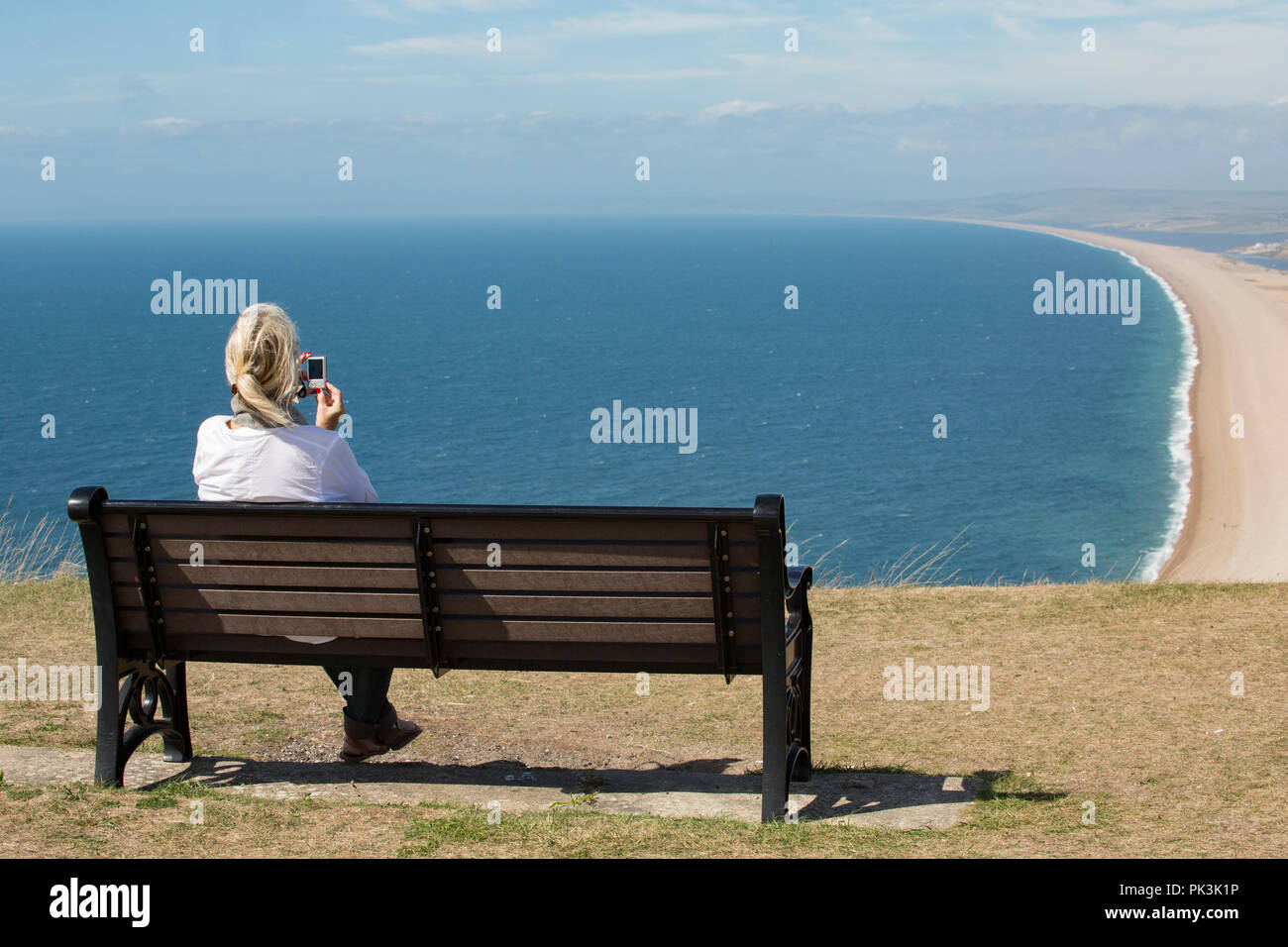 A woman taking a picture of Chesil beach from a bench situated on the Isle of Portland at the eastern end of Chesil beach. Dorset England UK GB Stock Photo