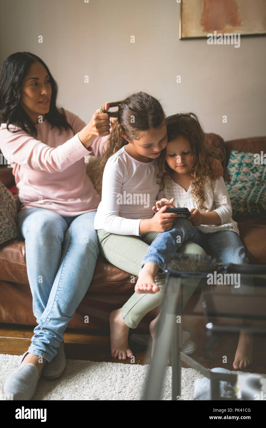 Daughter using mobile phone while mother making her hairstyle Stock Photo
