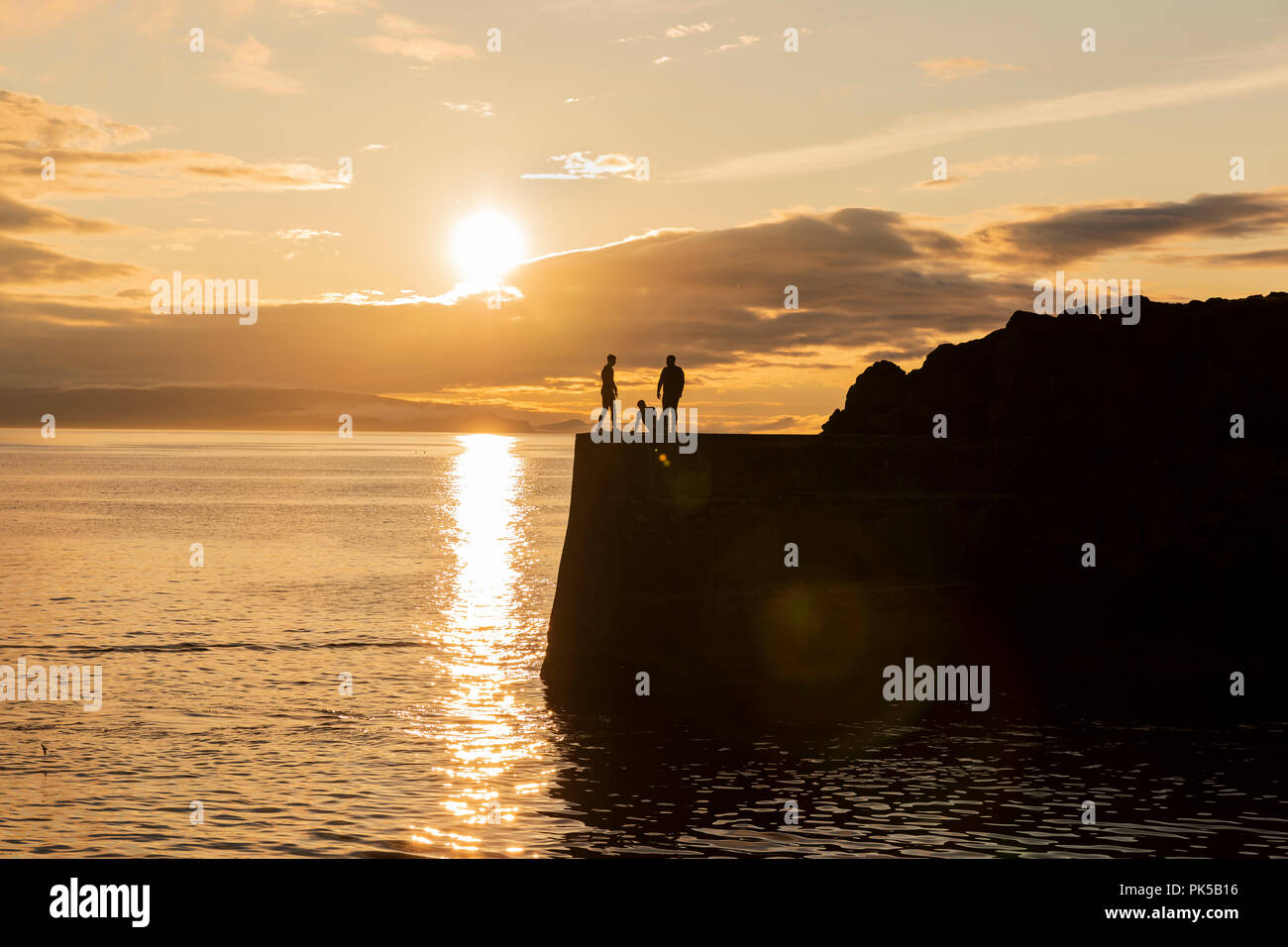 Silhouette of people jumping from the harbour wall at sunset into light of the setting sun reflected on sea, Portstewart, Co. Derry, Northern Ireland Stock Photo