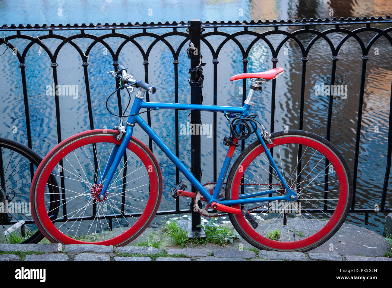 Bicycle with bright blue frame and red wheels leaning against railings beside a canal. Stock Photo