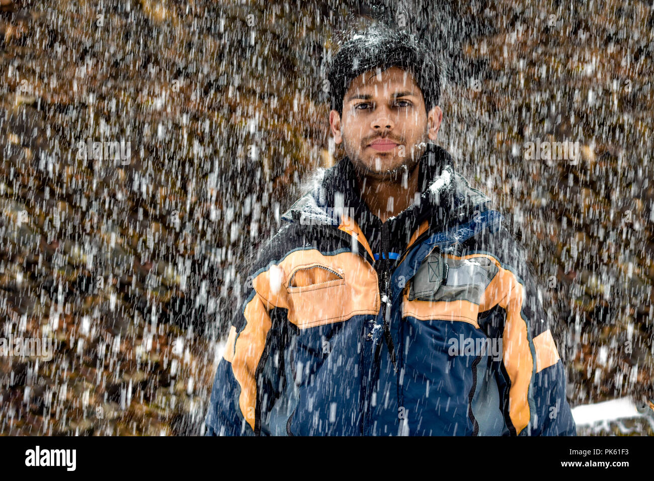 Young, Fair and Handsome Indian boy in early 20s in skiing costume, wearing snowsuit, standing still and posing in neutral mood during heavy snowfall Stock Photo