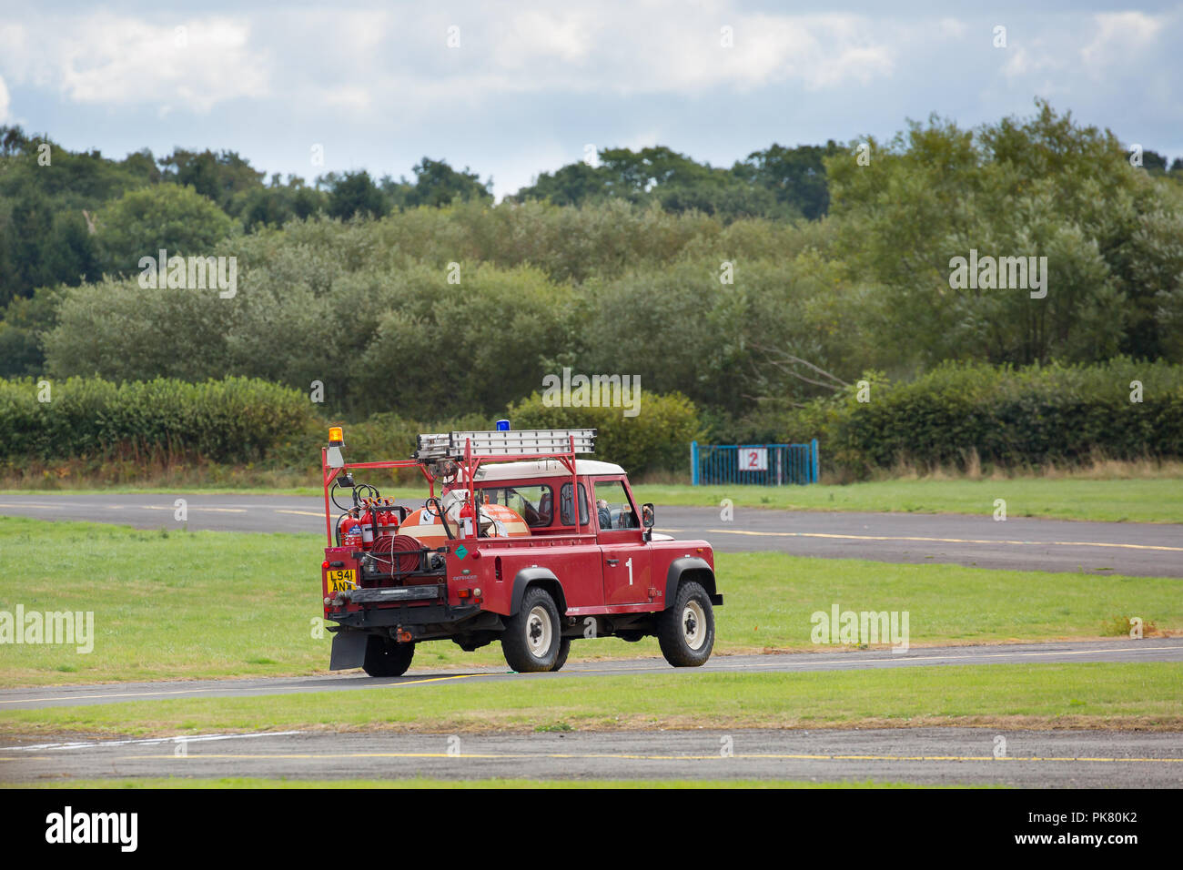 Emergency fire-fighting vehicle, blue light flashing, driving to runway at airfield to await landing of small light aircraft possibly in difficulty. Stock Photo