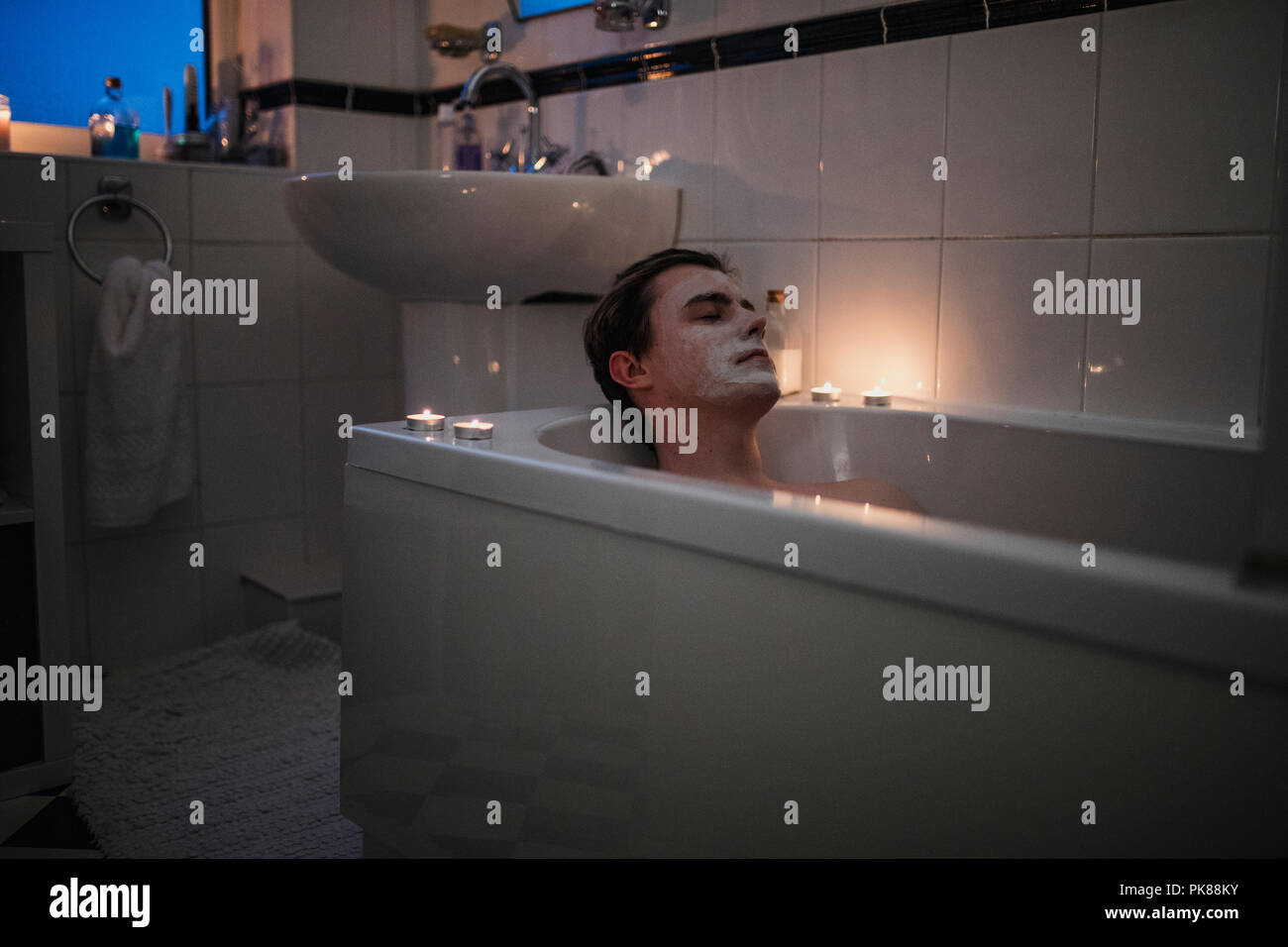 Young man is relaxing in the bath with a face mask on and candles lit around him. Stock Photo