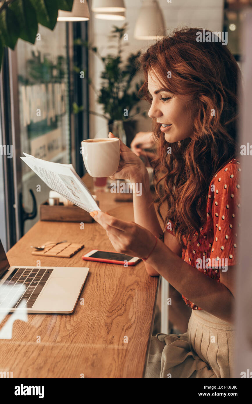 side view of woman with cup of coffee reading newspaper at table with laptop in coffee shop Stock Photo