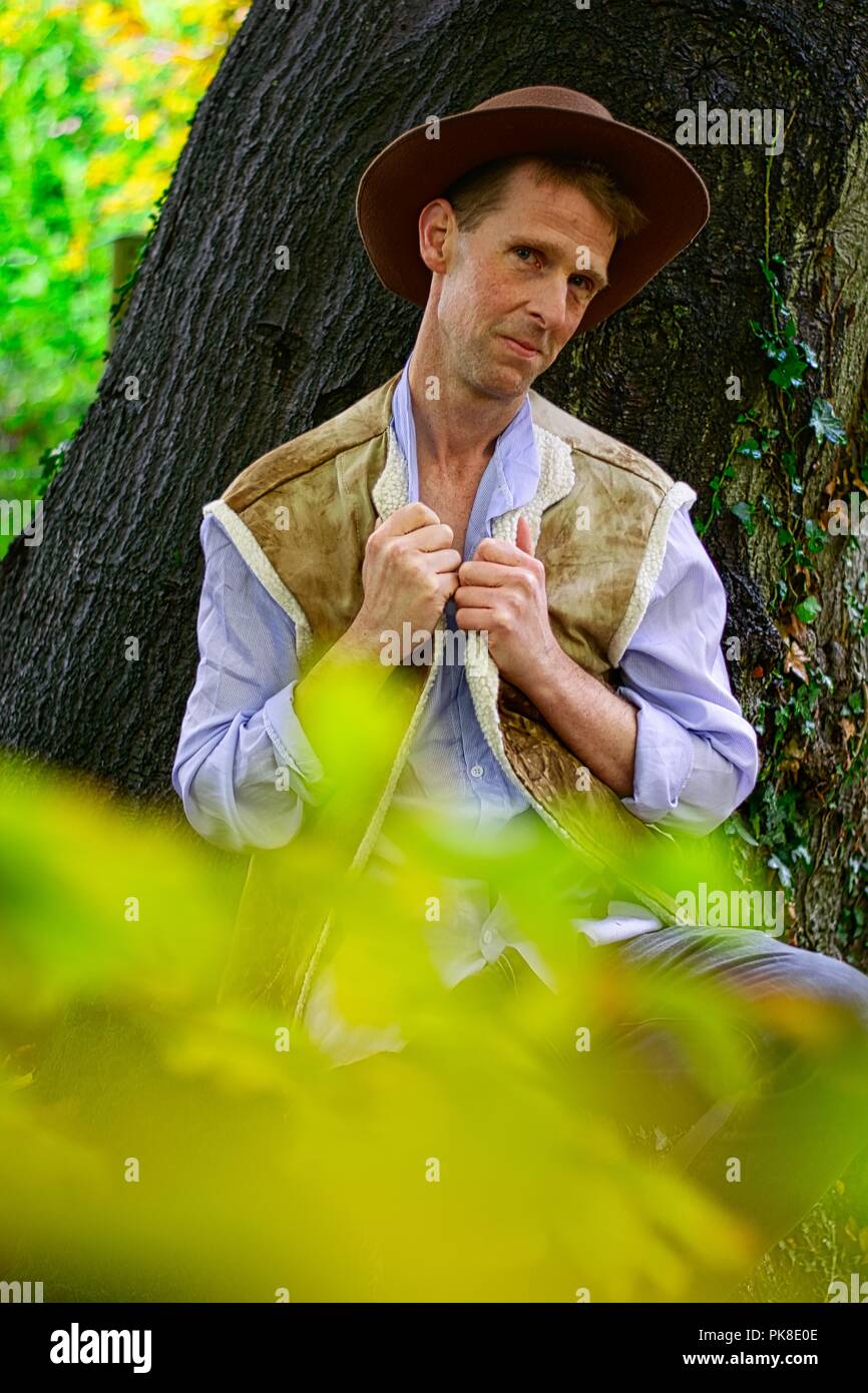 A man dressed like a cowboy leans against a tree trunk holding the collar of his shirt Stock Photo