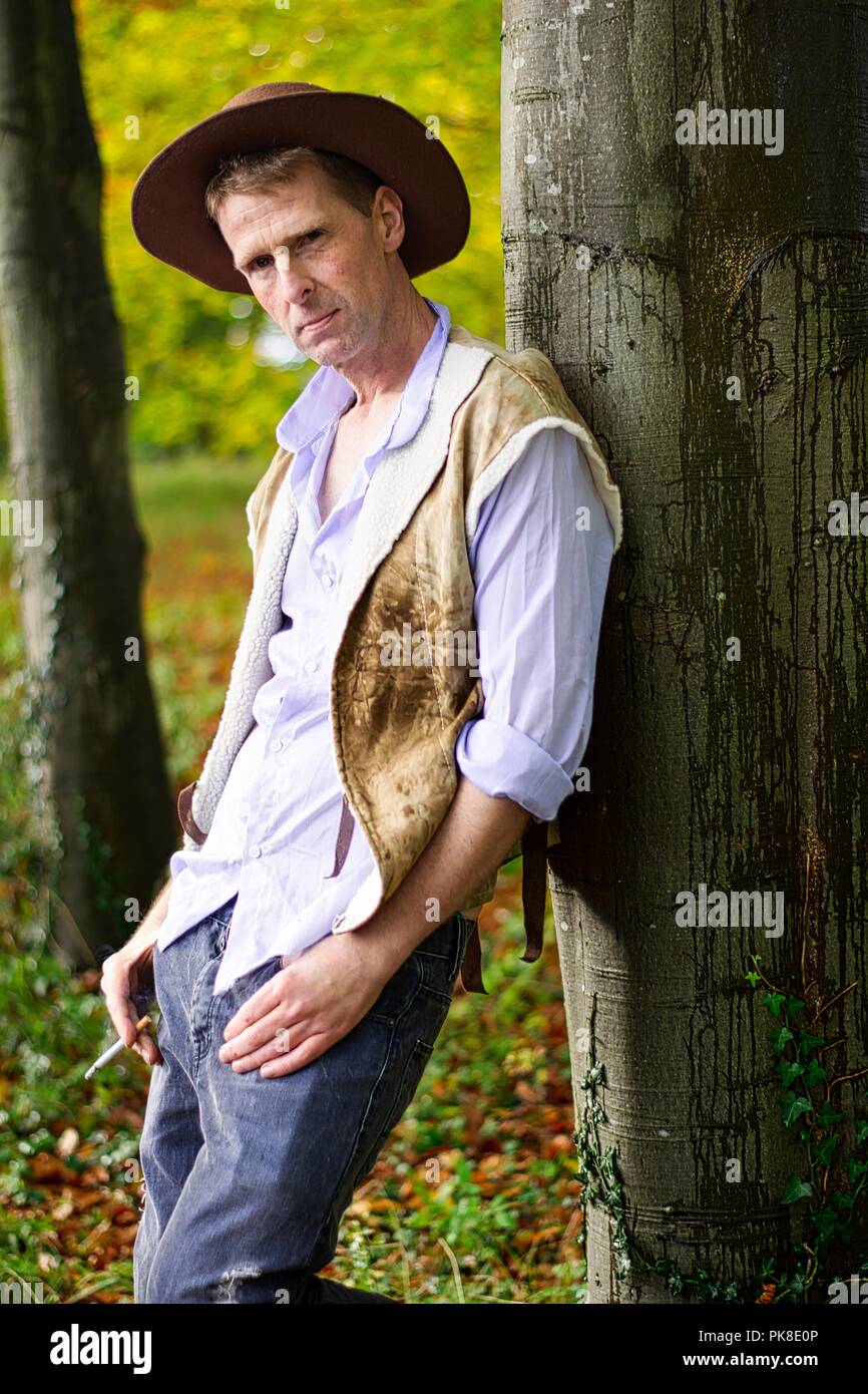 A man dressed like a  cowboy leans against a tree smoking a cigarette Stock Photo