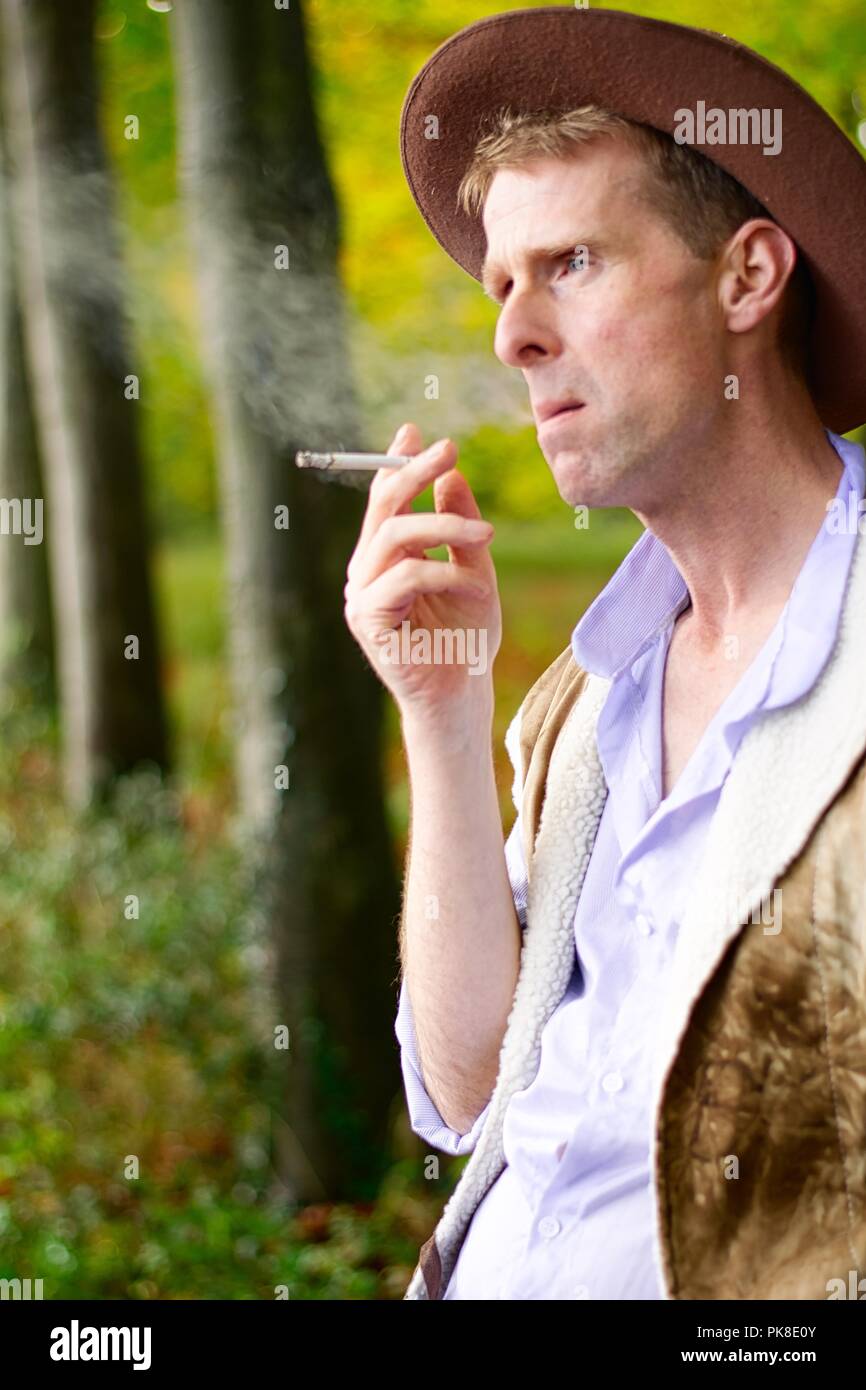 A man dressed like a  cowboy leans against a tree smoking a cigarette Stock Photo