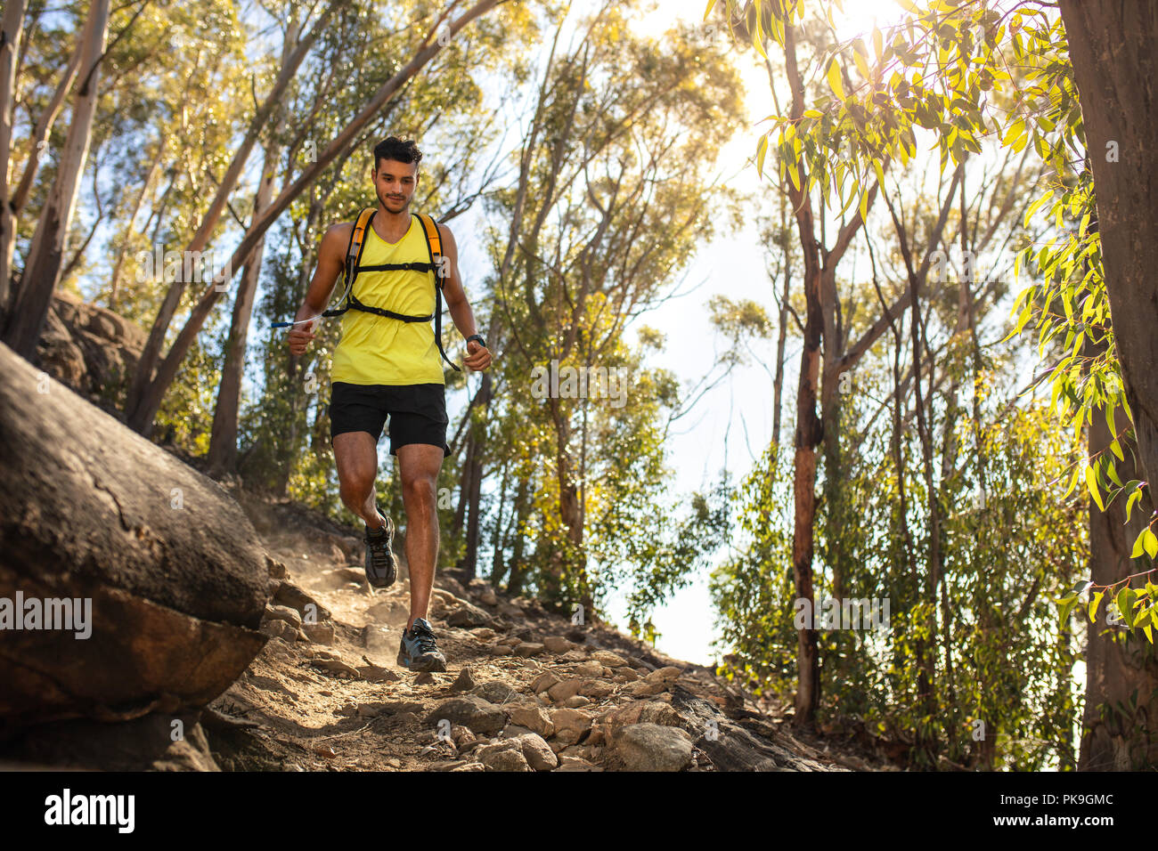 Fit young man in sportswear wearing hydration pack running outdoors over rocky trail on the hill. Mountain trail runner practicing on dirt path. Stock Photo
