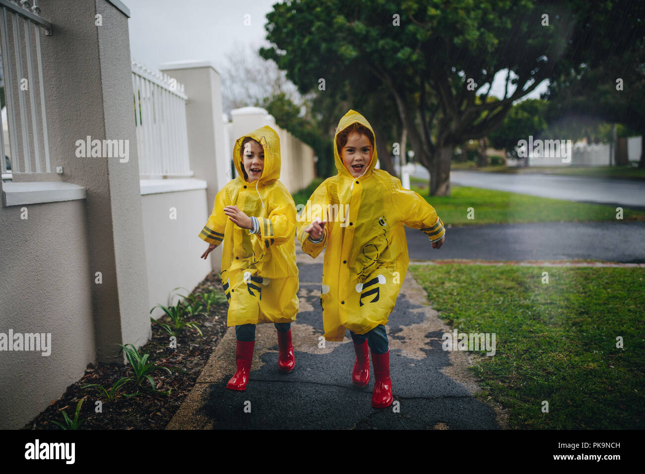 Happy young girls in raincoat and rubber boots running outdoors. Twin