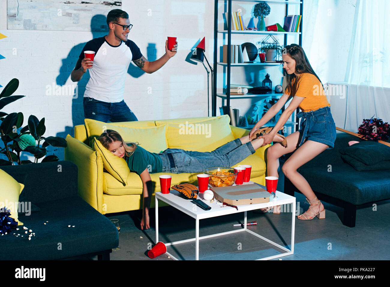 smiling man holding plastic cups and looking at girl pulling drunk female  friend sleeping on sofa after home party Stock Photo - Alamy