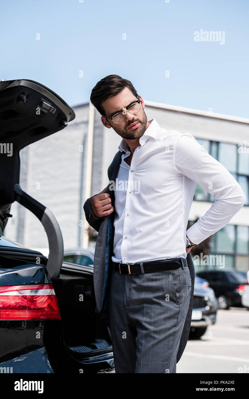 low angle view of handsome businessman taking off jacket and looking away near car Stock Photo