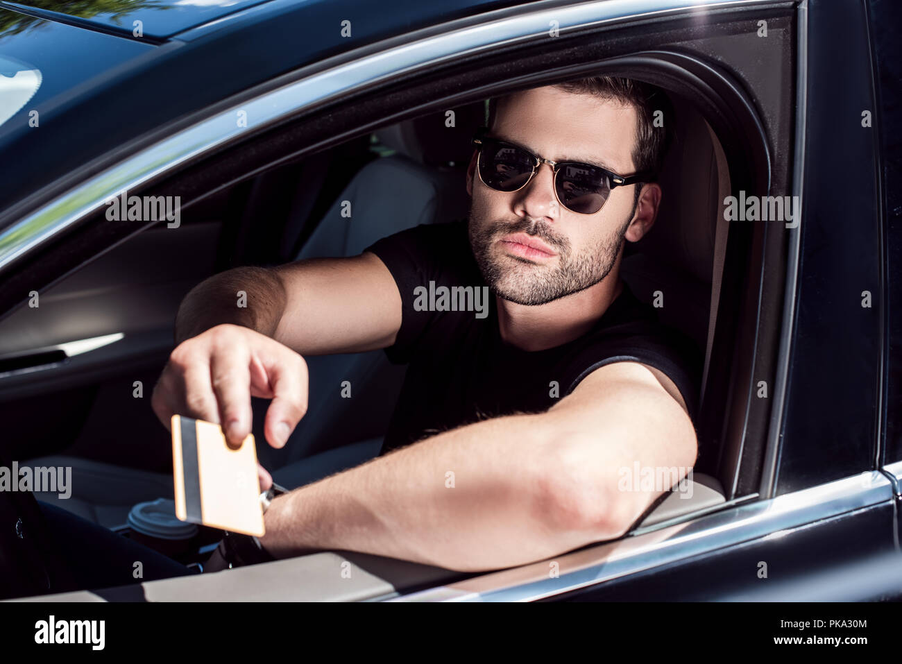 serious young man in sunglasses giving credit card while sitting in his car Stock Photo