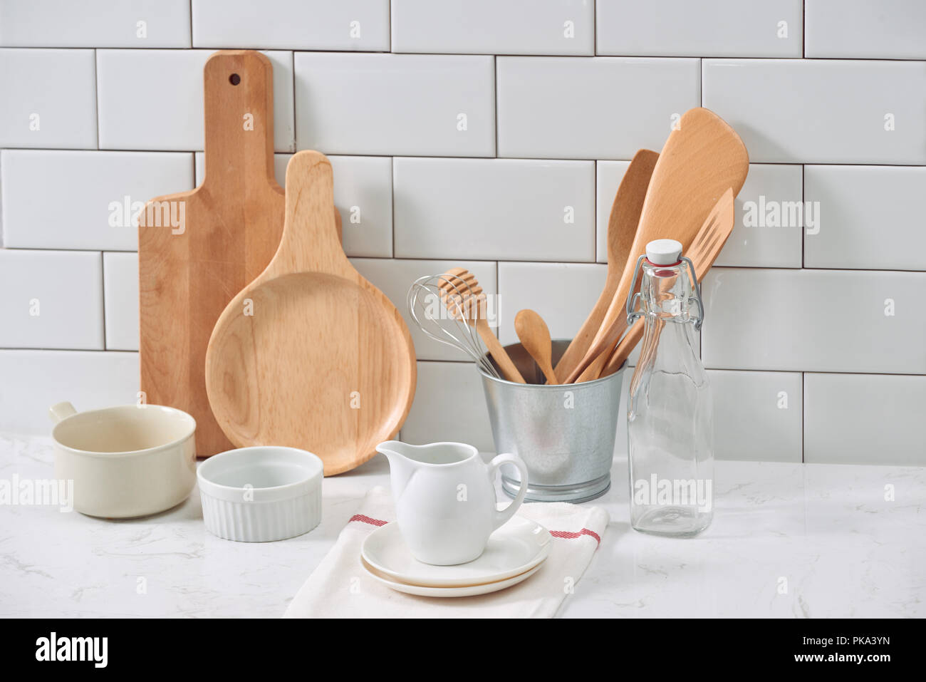 Simple rustic kitchenware against white wooden wall: rough ceramic pot with wooden cooking utensil set, stacks of ceramic bowls, jug and wooden trays. Stock Photo